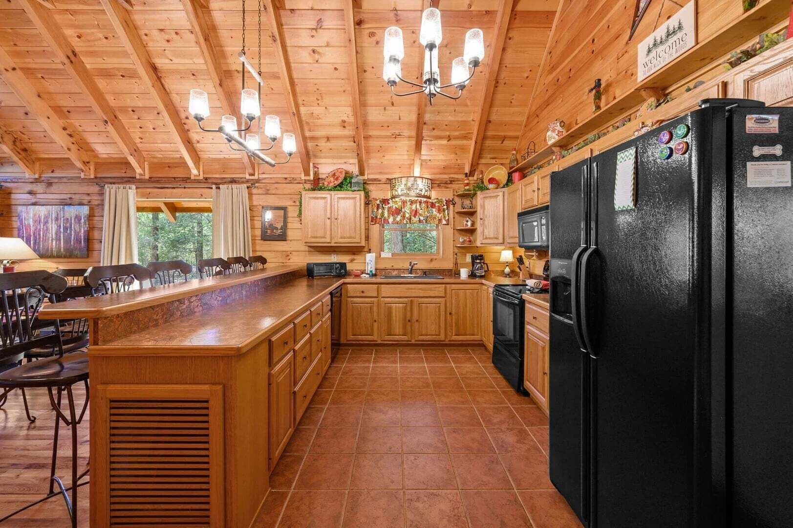 A wooden cabin interior with a kitchen featuring a black refrigerator, wooden cabinets, and a central island with chairs.