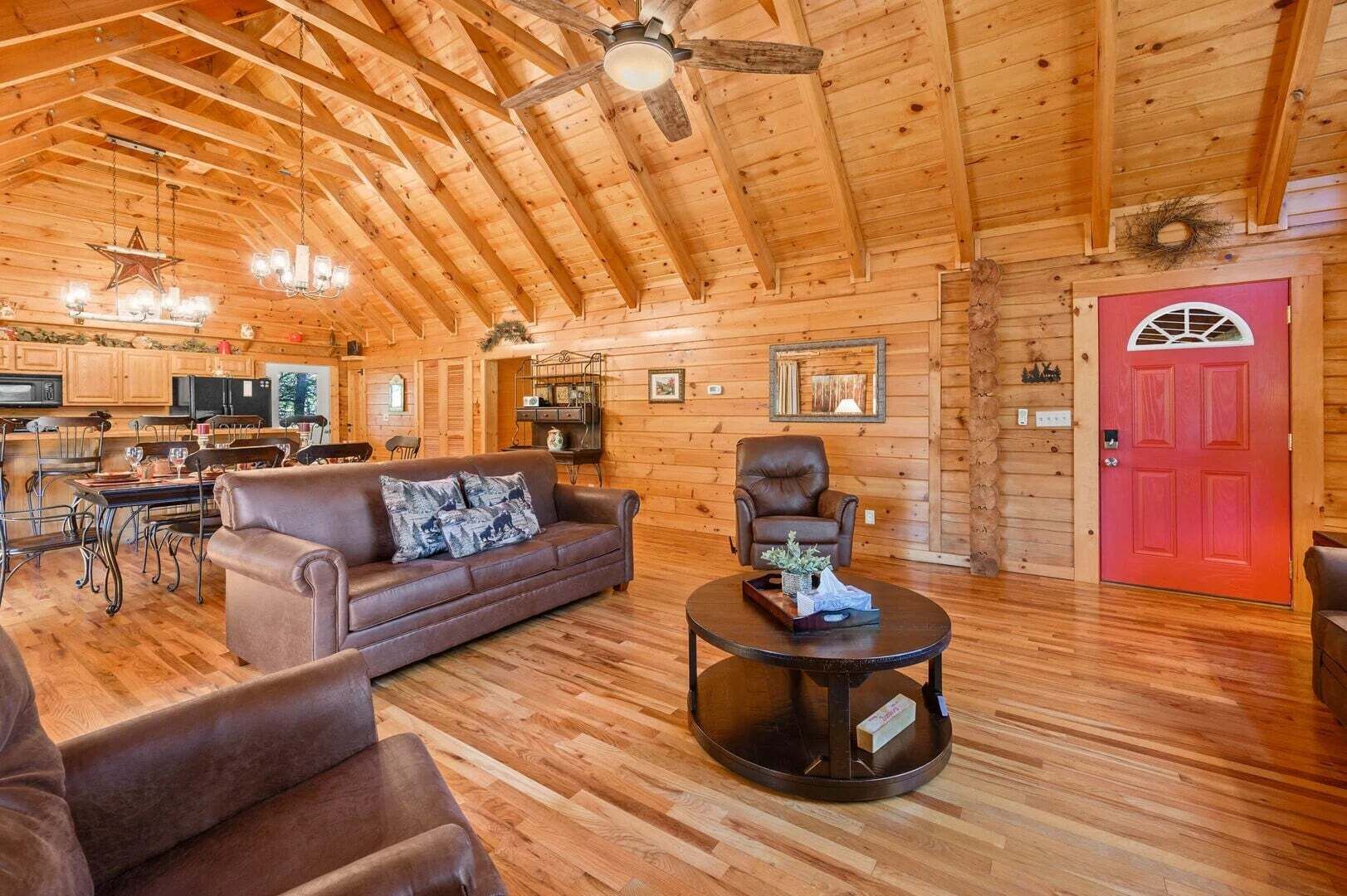 Interior de una cabaña de madera con comedor, sala de estar con sofá y sillones, una puerta roja y un ventilador de techo.