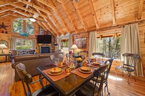 Interior de una cabaña de madera con comedor, sofá, chimenea y ventanas amplias.