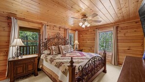 Un dormitorio de cabaña de madera con una cama, dos mesitas de noche, un ventilador de techo y ventanas grandes.
