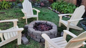 Un pozo de fuego circular rodeado por cuatro sillas Adirondack en un jardín con césped, una valla de madera y algunas plantas al fondo.