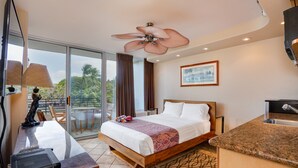 Ein Hotelzimmer mit Bett, Schreibtisch mit Computer, Deckenventilator, Fenster mit Vorhängen und Blick auf Palmen.