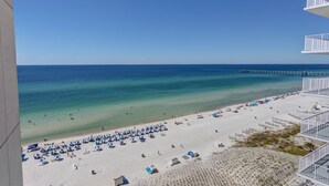 Ein Strand mit Sonnenschirmen und Menschen, ein Steg, der sich ins Wasser erstreckt, sowie ein Gebäude mit Balkonen.