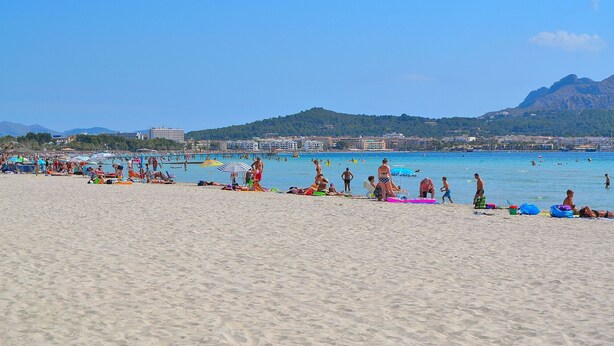 A beach with people sunbathing and swimming, a clear blue sky, and mountains in the background.