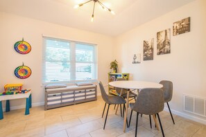 A dining area with a round table and four chairs, a window with blinds, a bookshelf, and a colorful toy table.