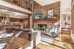 A spacious living area with a stone fireplace, a kitchen with wooden cabinets, and a high ceiling with a wooden balcony. - Charlie Rentz: Bachelor Gulch Ski-In/Out, Sleeps 12, Ritz Pool + Hot Tub Access, 2 King Suites (Avon)