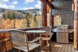 Una terraza de madera con sillas de mimbre, una mesa, una parrilla y vistas a árboles otoñales y colinas.