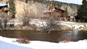Un chalet rustique aux murs en bois, situé au bord d’une rivière où l’on aperçoit encore des plaques de neige, entouré d’arbres et adossé à des collines.