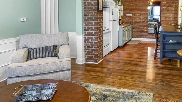 A living room with a sofa, a coffee table, and a kitchen area with a brick wall and white appliances.