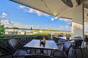 Un patio exterior con mesas y sillas, con vista a una playa y una estructura azul distante.