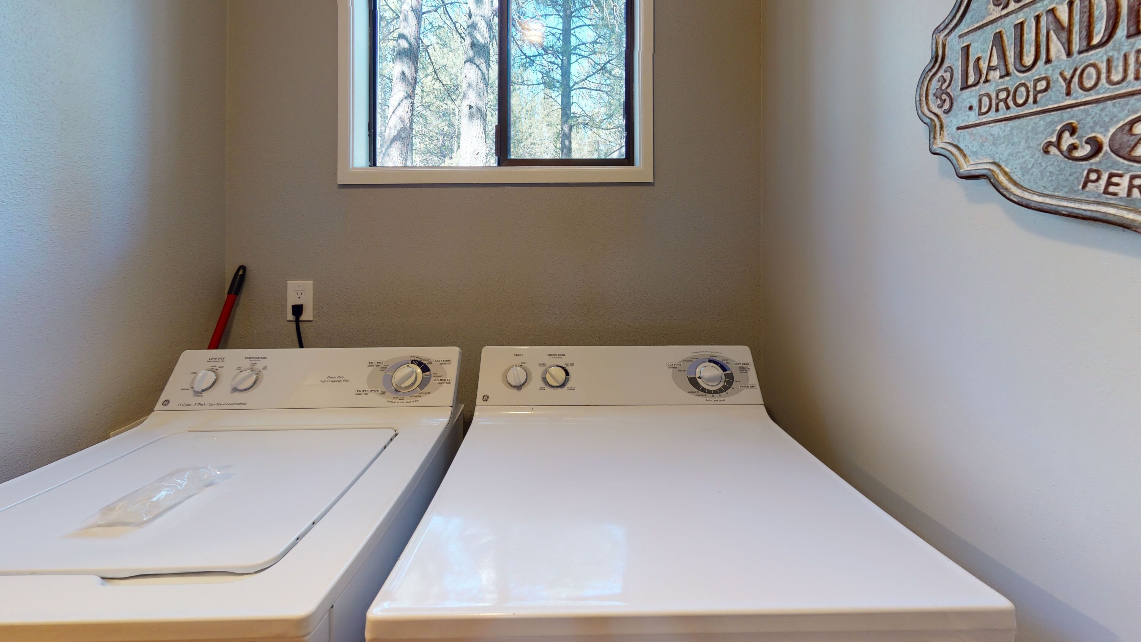 A laundry room with two front-loading washers and dryers, a window with a view of trees, and a wall-mounted sign.