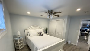 Un dormitorio con una cama blanca, una mesita de noche con una lámpara, un ventilador de techo y vistas a la cocina.