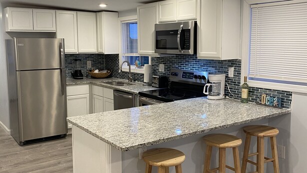 Una cocina moderna con armarios blancos, electrodomésticos de acero inoxidable, encimera de granito y taburetes de madera.