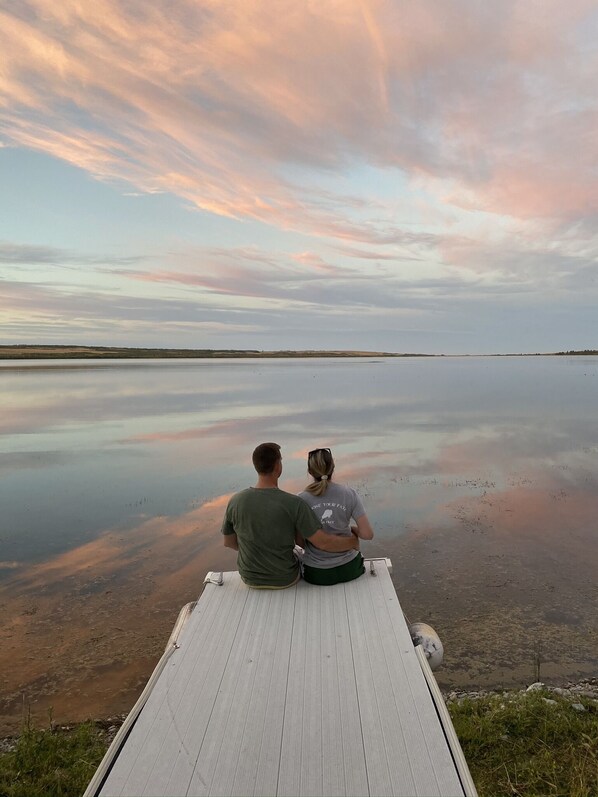 Two people sitting on a wooden bench overlooking a calm body of water with a sunset sky. - Spectacular Views! Minutes from Many Glacier and St. Mary's! (Babb)