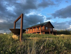 A rustic log cabin with a balcony, surrounded by tall grass and a cloudy sky. - Spectacular Views! Minutes from Many Glacier and St. Mary's! (Babb)