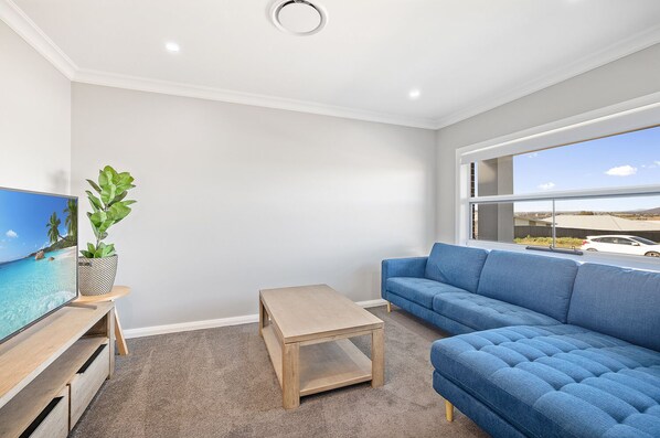 A modern living room with a blue sofa, a small wooden table, a TV on a wooden stand, and a potted plant. - Devan House - Your Innkeeper Mudgee (Mudgee)