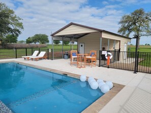 A pool area with lounge chairs, a covered seating area, and a building with a door and windows. - The Ranch Motel. Large cottage in a country setting with pool and hot tub. (Round Top)