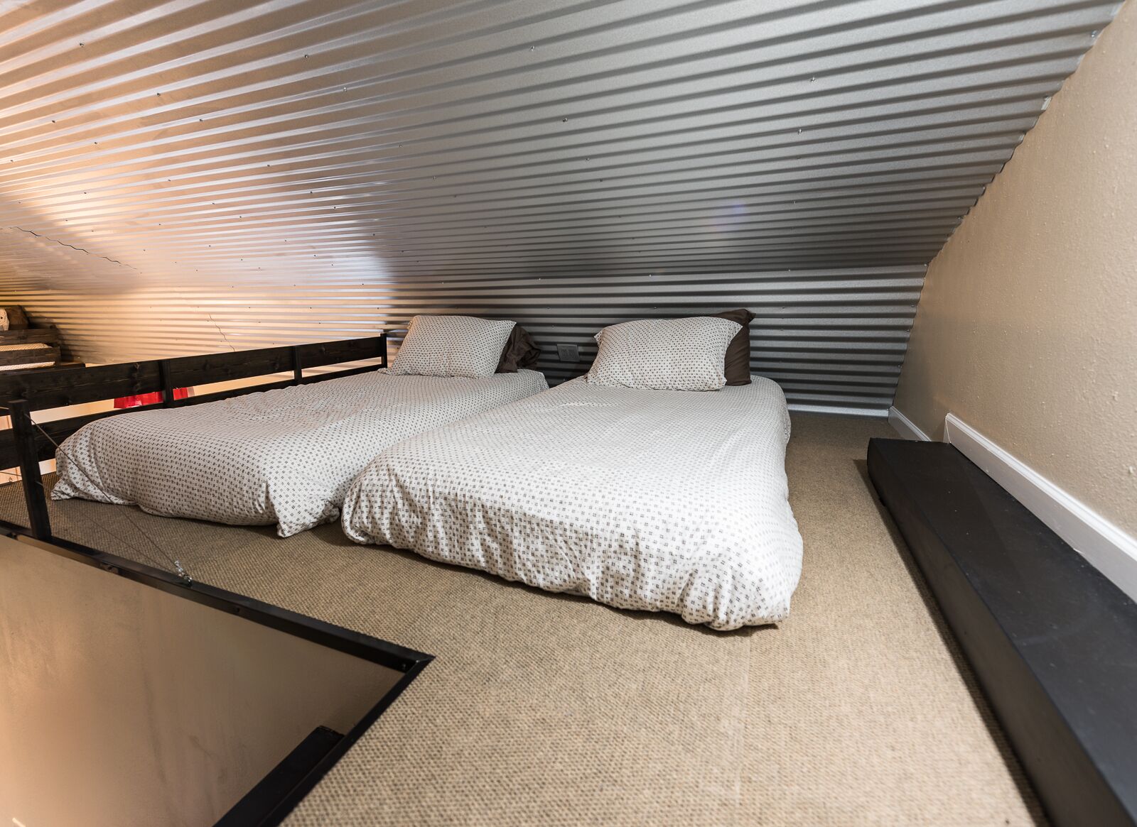 A compact bedroom with a sloped ceiling, two beds with matching bedding, and a small bedside table.
