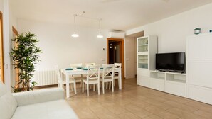 Un soggiorno moderno con un divano bianco, un tavolo da pranzo con sedie, una TV su un mobile bianco e una pianta grande.