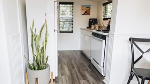 Una cocina estrecha con un refrigerador blanco, una estufa y un lavamanos. Hay una planta en maceta sobre un pedestal de madera y una silla negra.