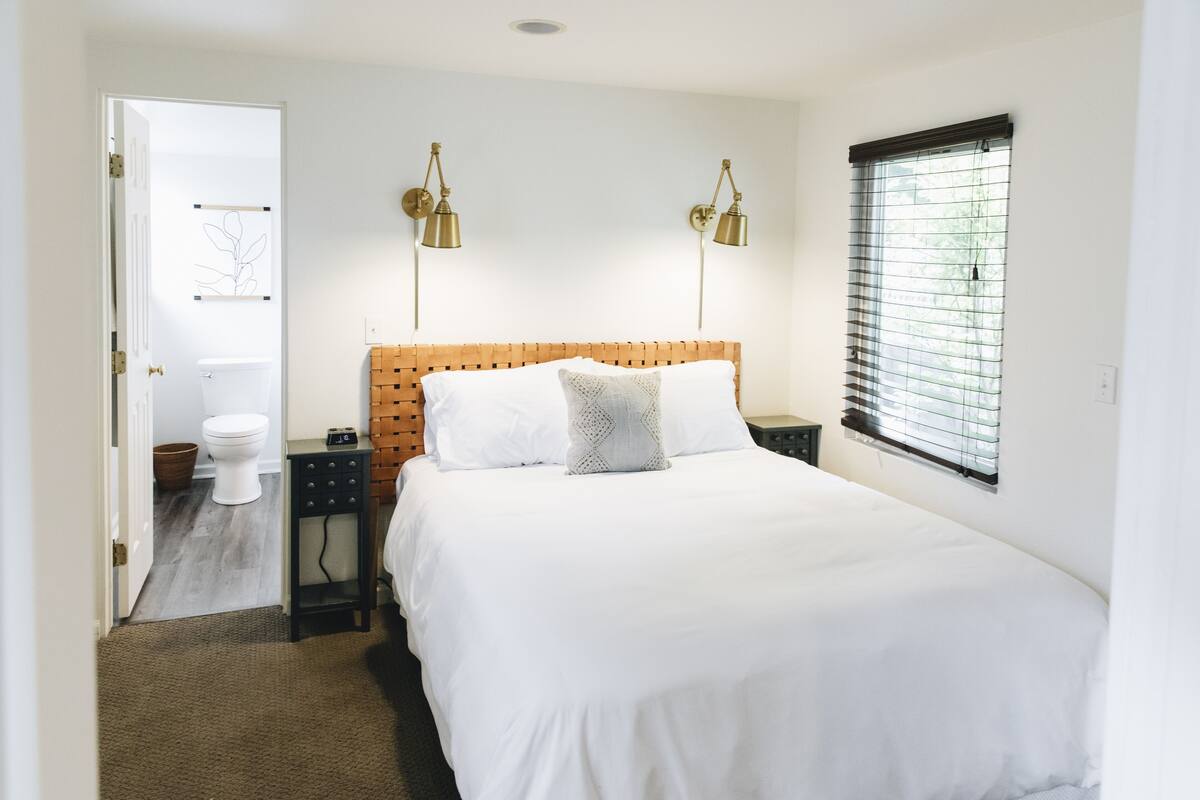 A bedroom with a wooden headboard, a large bed, two bedside lamps, a window with blinds, and a door leading to a bathroom.