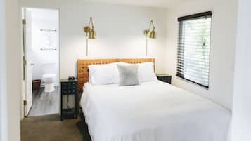 A bedroom with a wooden headboard, a large bed, two bedside lamps, a window with blinds, and a door leading to a bathroom.