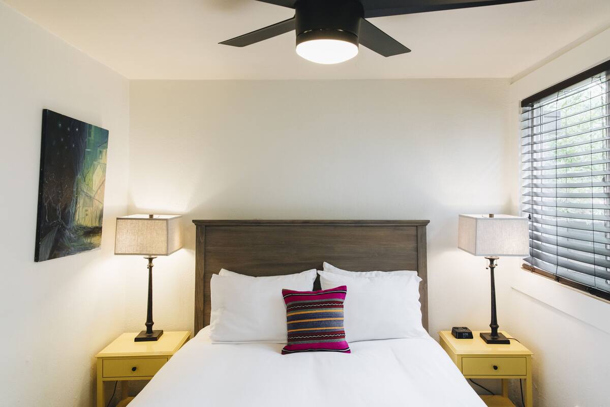 A neatly made bed with white linens, a colorful striped pillow, two bedside lamps, and a ceiling fan.