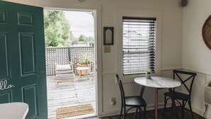 Una habitación con puerta verde, una mesita con dos sillas, una ventana con persianas y vista a un patio exterior con sillas y plantas.