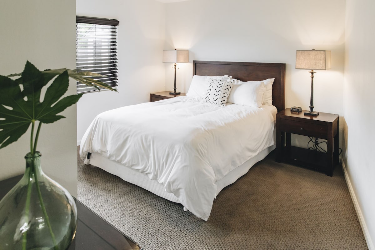 A neatly made bed with white linens, two bedside tables with lamps, and a large plant in a vase.