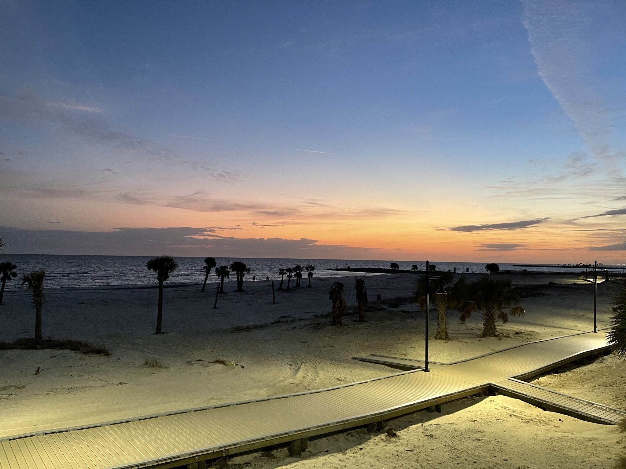 Uma praia com um calçadão de madeira que leva à água, palmeiras e um pôr do sol ao fundo.