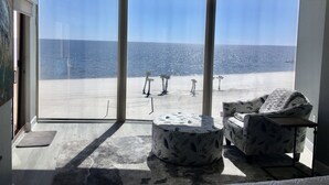 Ein Zimmer mit einem großen Schiebeglastür, die Blick auf einen Strand bietet. Im Inneren befindet sich ein Muster-Hocker und ein Sessel mit gemustertem Bezug.