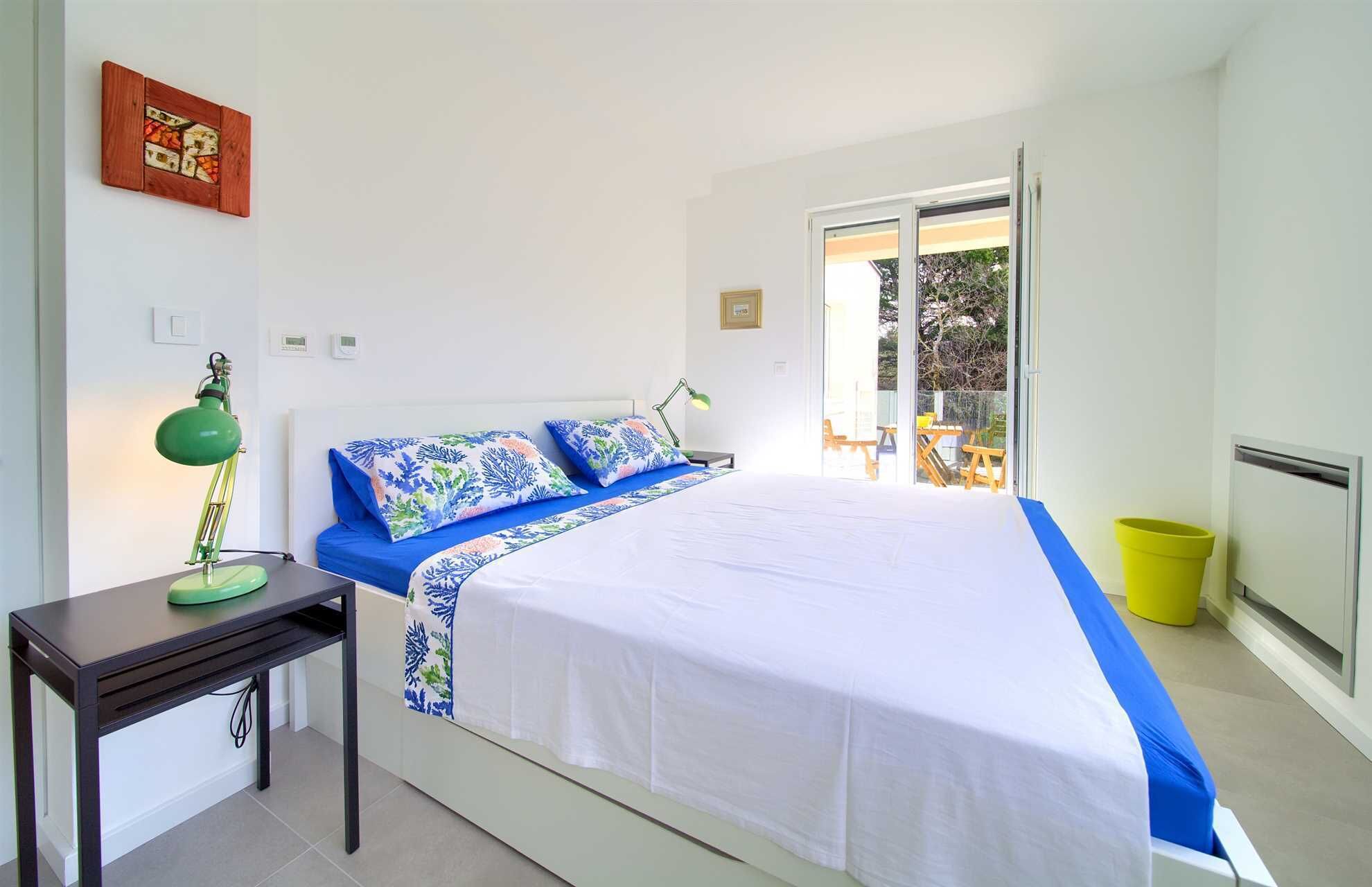 A bedroom with a bed, a nightstand, a desk, a window, and a door leading to a balcony.