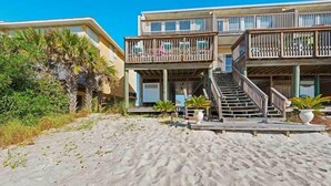 Un hotel frente a la playa con una terraza de madera, una escalera que conduce al nivel superior y una palmera en primer plano.