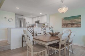 A dining area with a wooden table and chairs, a chandelier, and a kitchen in the background.