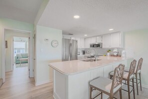 A modern kitchen with white cabinets, a central island with bar stools, stainless steel appliances, and a visible clock.