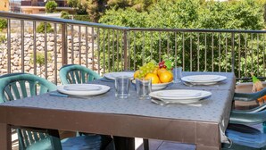 Un área de comedor al aire libre con una mesa lista para dos personas, rodeada de barandas metálicas, con vistas a edificios y árboles en el fondo.