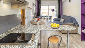 Una cocina compacta con estufa integrada, un comedor con mesa y sillas, y una sala de estar con sofá y una estantería morada.