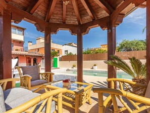 Un área de estar al aire libre con techo, mobiliario de mimbre, piscina y vistas a edificios residenciales.