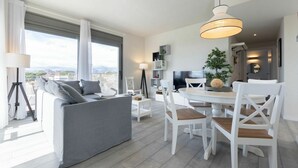 A modern living room with a grey sofa, white dining table, and chairs, a TV, and a large window with curtains. - Beautiful apartment for 4 guests with WIFI, pool, A/C, TV and terrace (Cambrils)