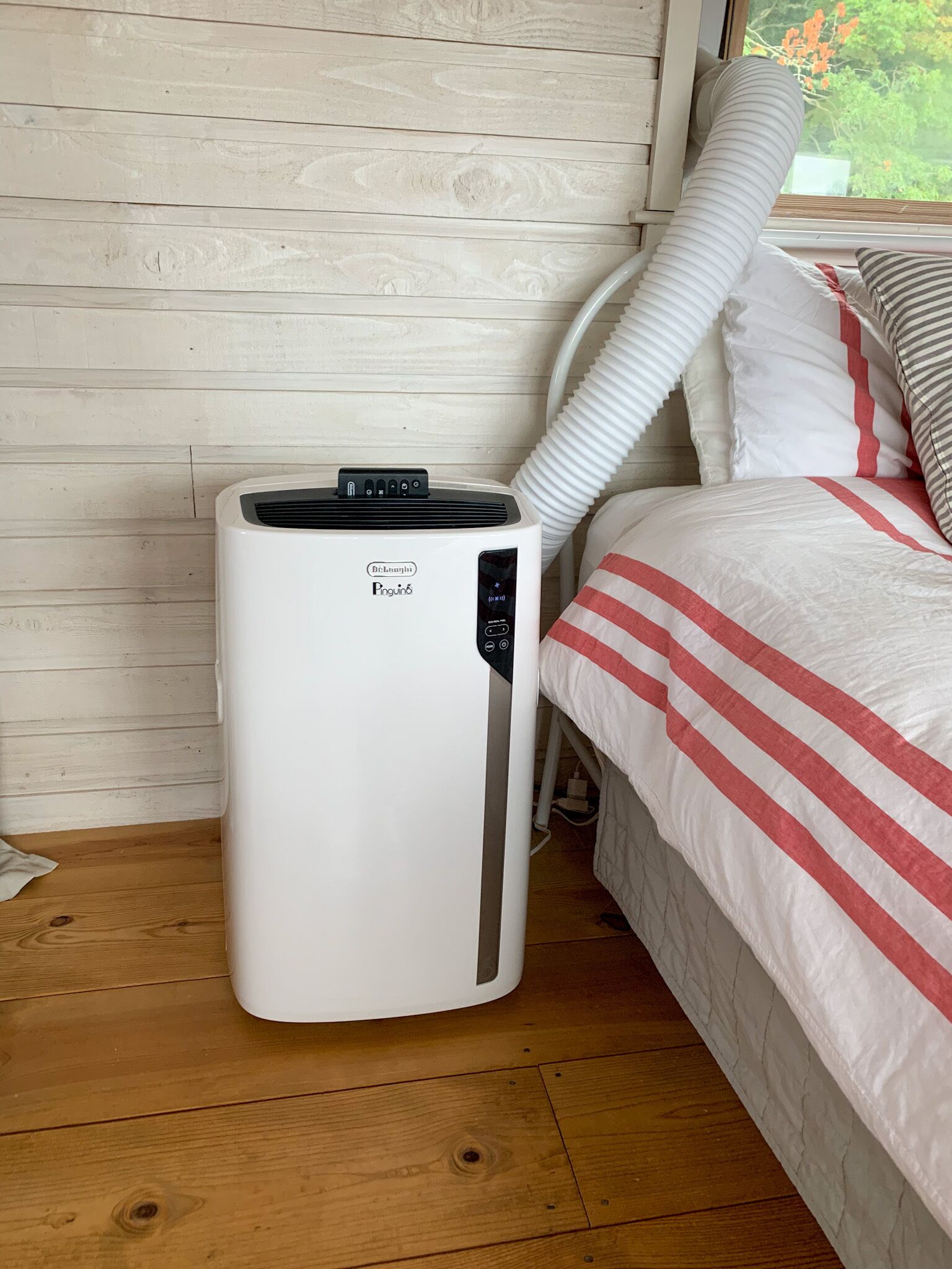 A portable air conditioner in a room with a wooden floor and a bed with striped bedding.