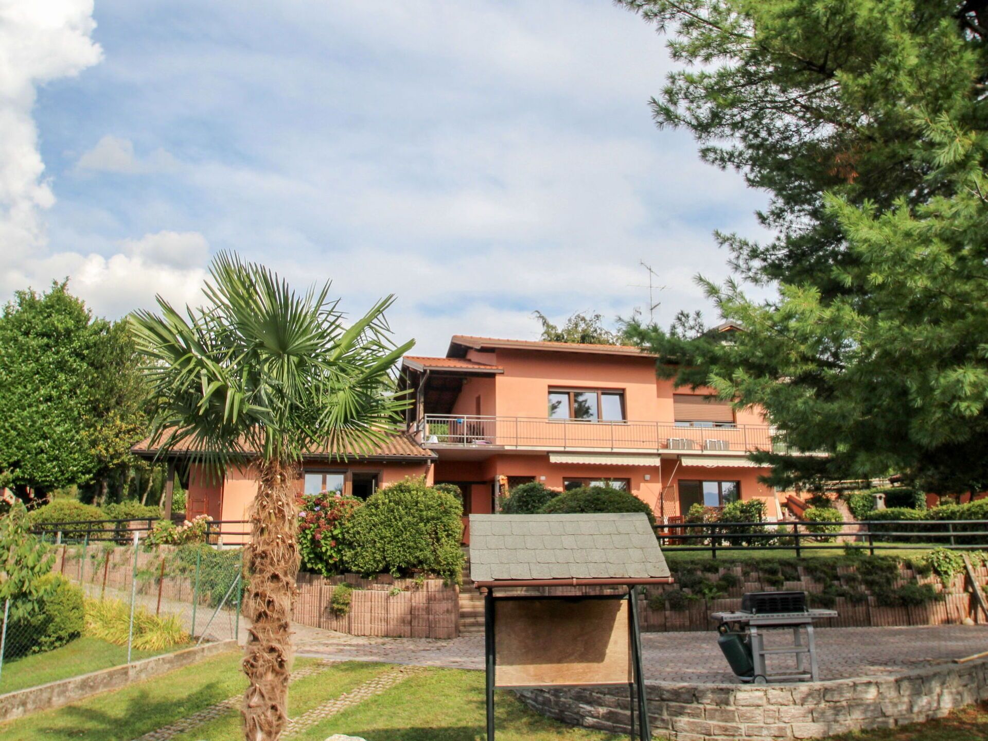 A two-story building with a balcony, surrounded by greenery and a small fenced area.