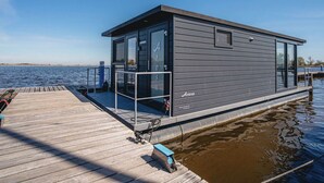 Una moderna casa galleggiante con terrazza in legno, porte in vetro e il nome 'Arosa' sul fianco.