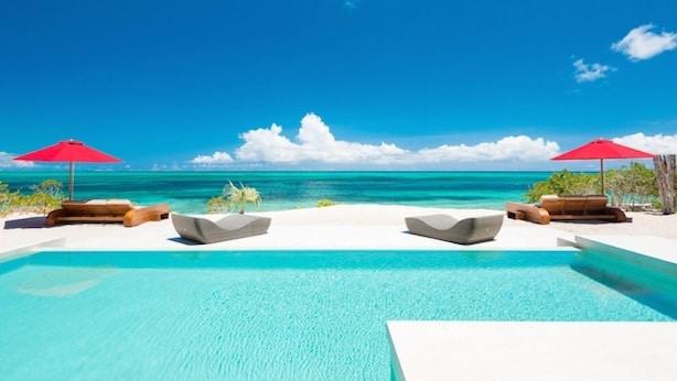 A poolside area with lounge chairs and red umbrellas, overlooking a clear blue sea and a white sandy beach. - Beach Kandi 15% off stays in Fall 2026 | Beach Front - Located in  Magnificent Grace Bay with House Cleaning Included (Providenciales)