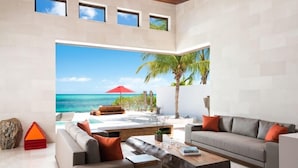 A modern living room with a large window overlooking a beach and ocean. - Beach Kandi 15% off stays in Fall 2026 | Beach Front - Located in  Magnificent Grace Bay with House Cleaning Included (Providenciales)
