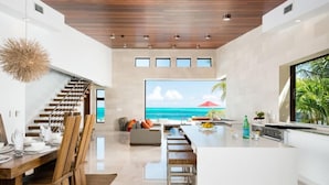 A modern kitchen with a large island, a dining area with wooden chairs, and a view of the ocean through a large window. - Beach Kandi 15% off stays in Fall 2026 | Beach Front - Located in  Magnificent Grace Bay with House Cleaning Included (Providenciales)