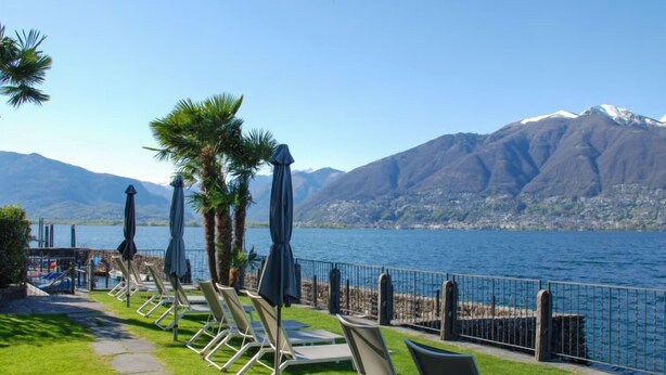 Eine Seeterrasse mit Liegestühlen und Sonnenschirmen mit Blick auf einen Berg und See.