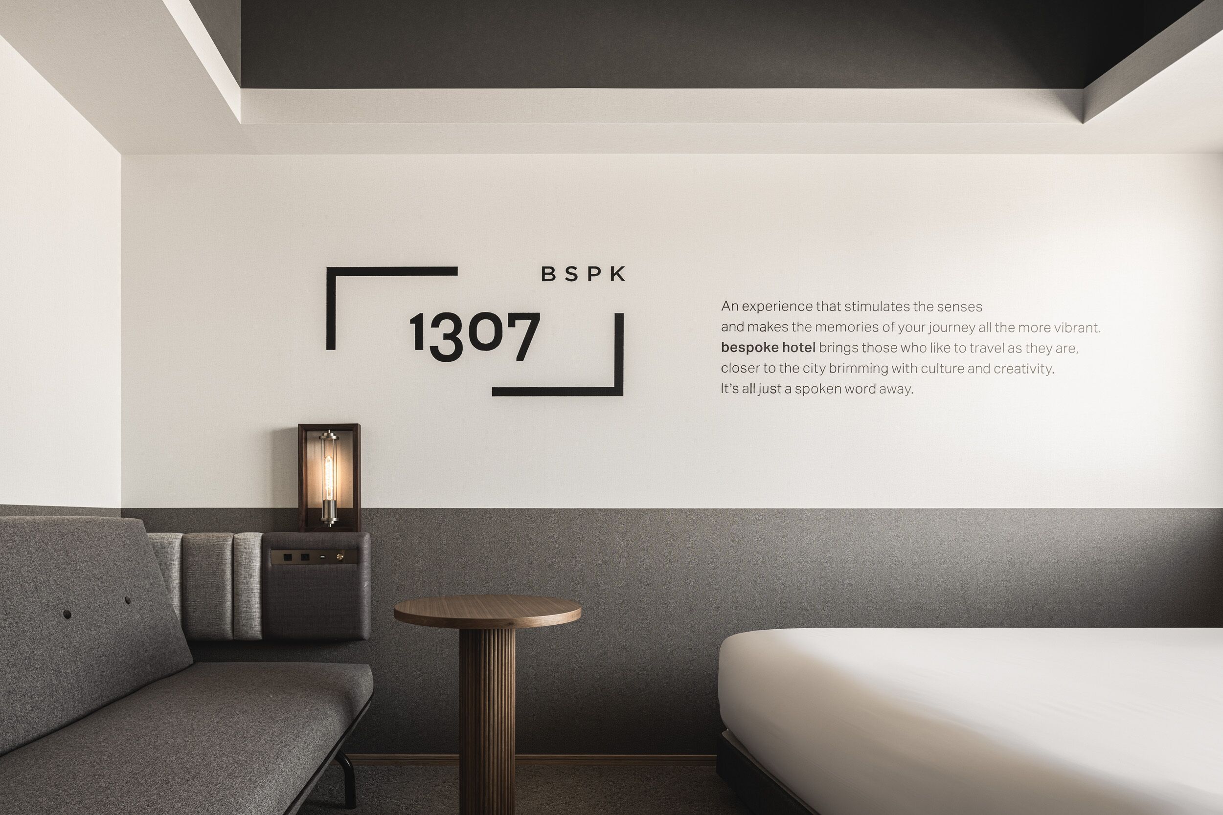 A modern hotel room with a grey sofa, a small round table, a bedside lamp, and a bed. The wall features a large number '1307' and the text 'BSPK' and a description of the hotel experience.