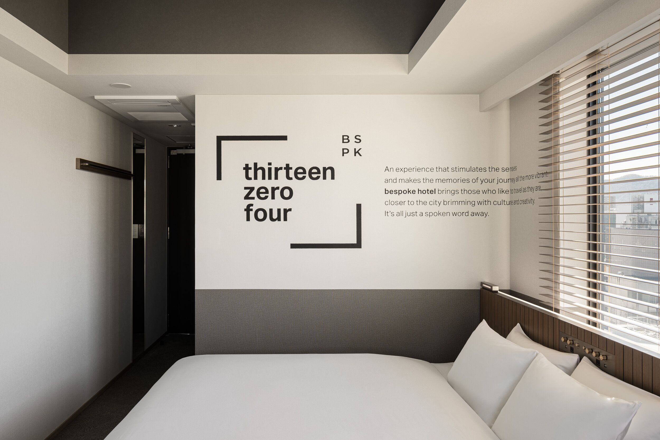 A modern hotel room with a large bed, a view of the city through floor-to-ceiling windows, and a wall-mounted sign with the text "thirteen zero four".