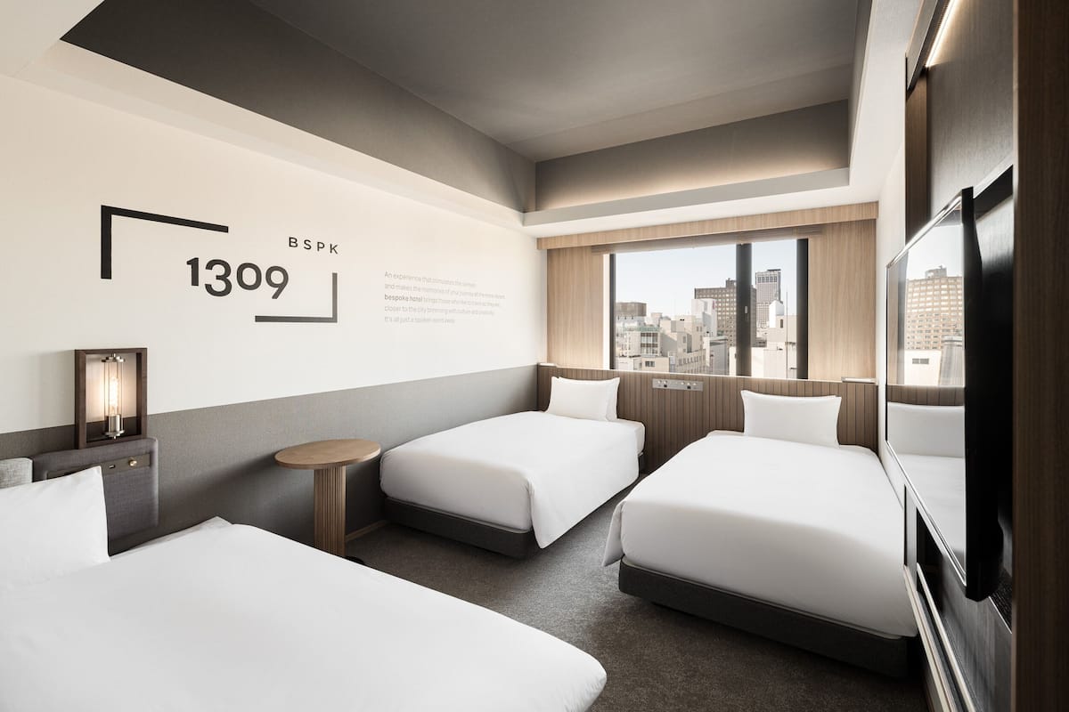 A hotel room with two beds, a small table, a lamp, and a large window offering a city view.