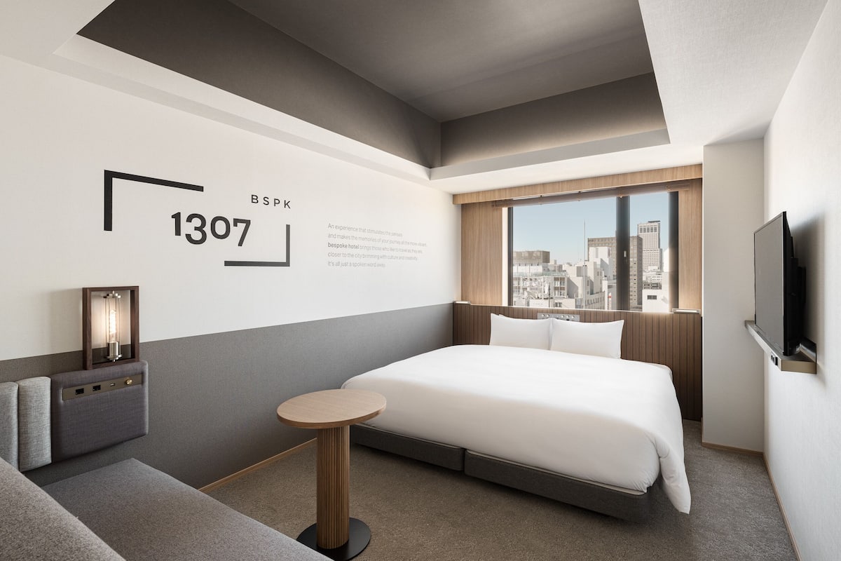 A modern hotel room with a large bed, a small round table, a TV, and a city view.